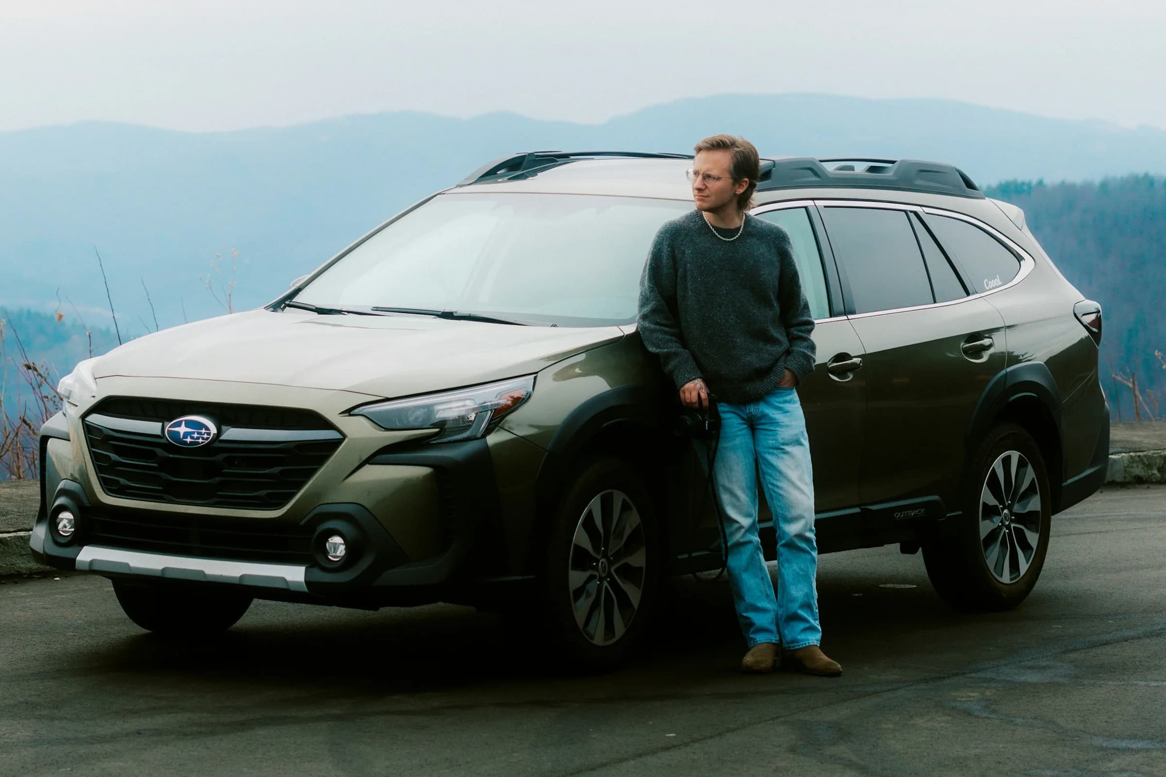 Man in a gray sweater leaning against a green Subaru SUV at a misty mountain overlook