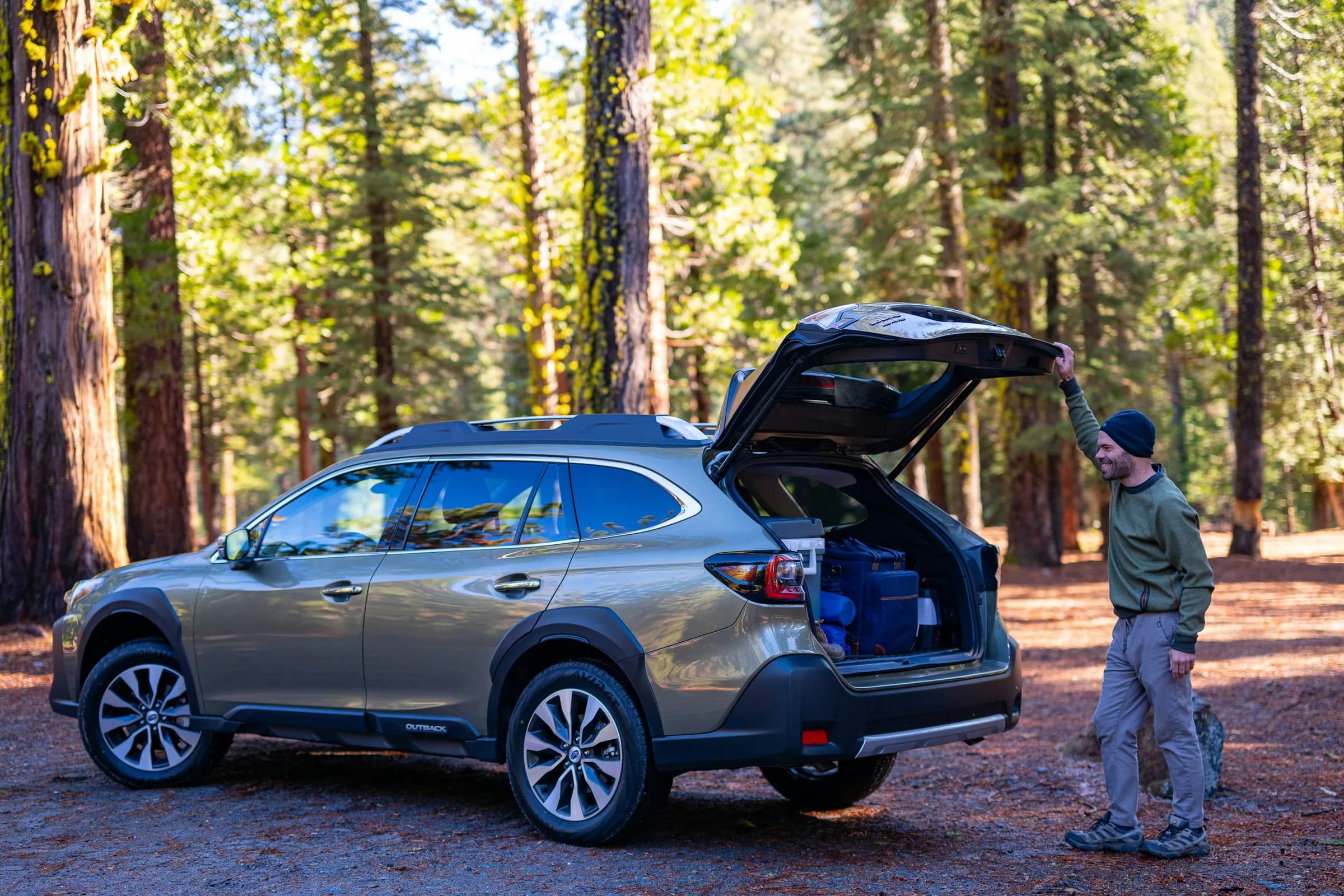A man lifts the open hatch of a Subaru SUV packed with camping gear in a sunlit pine forest