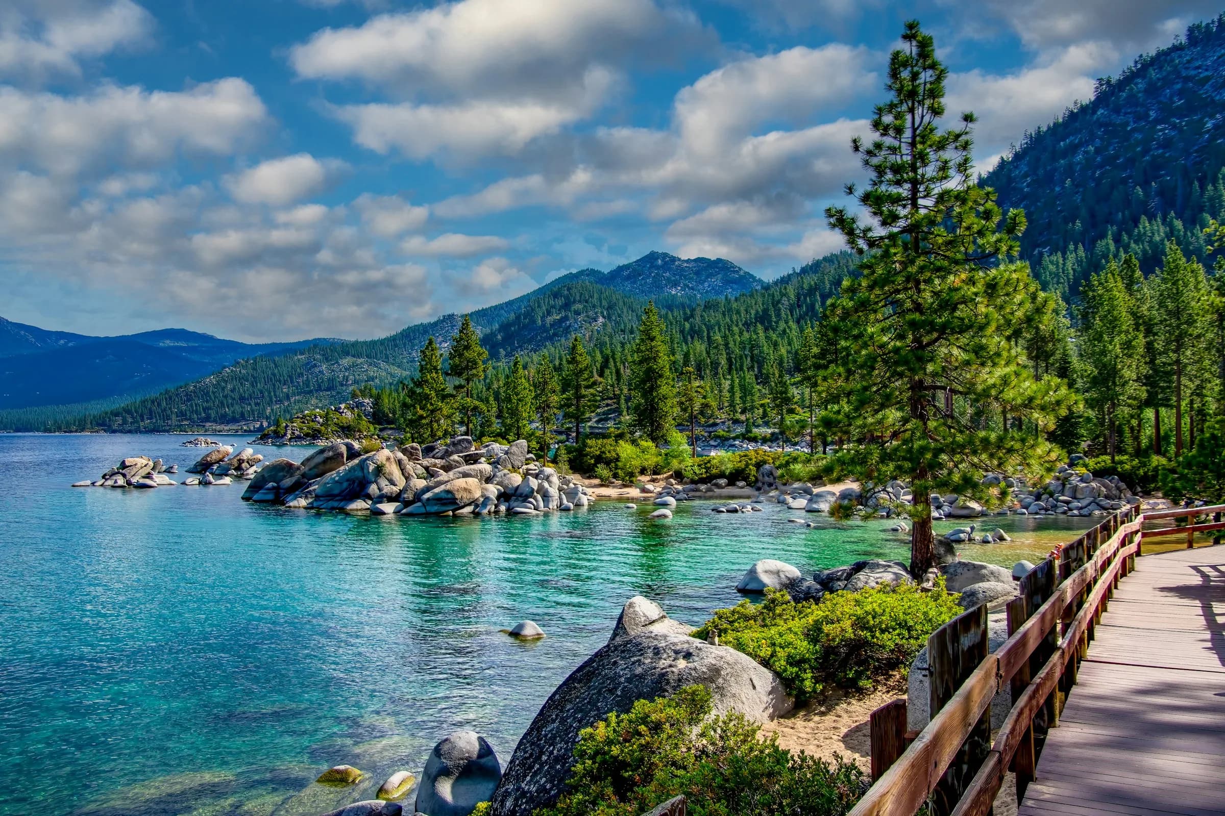 Turquoise water and granite boulders beside a lakeside boardwalk on Lake Tahoe, with pines and forested mountains beyond