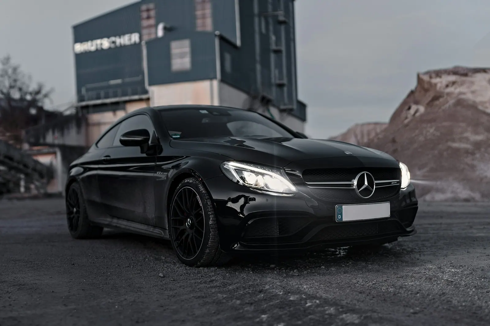 Black luxury coupe with its headlights on, parked on a gravel lot