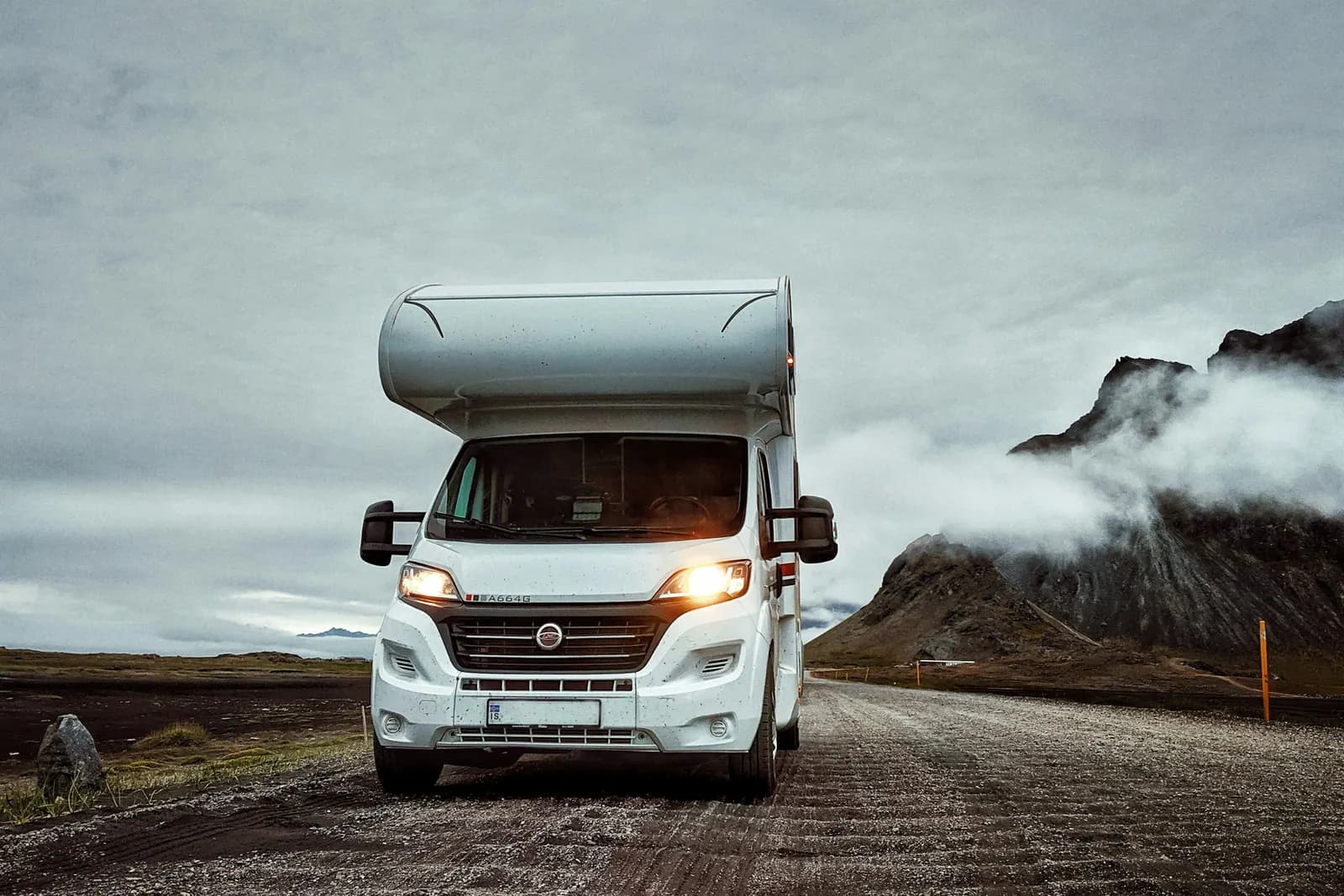 White Class C motorhome with headlights on, driving a mountain road under low clouds