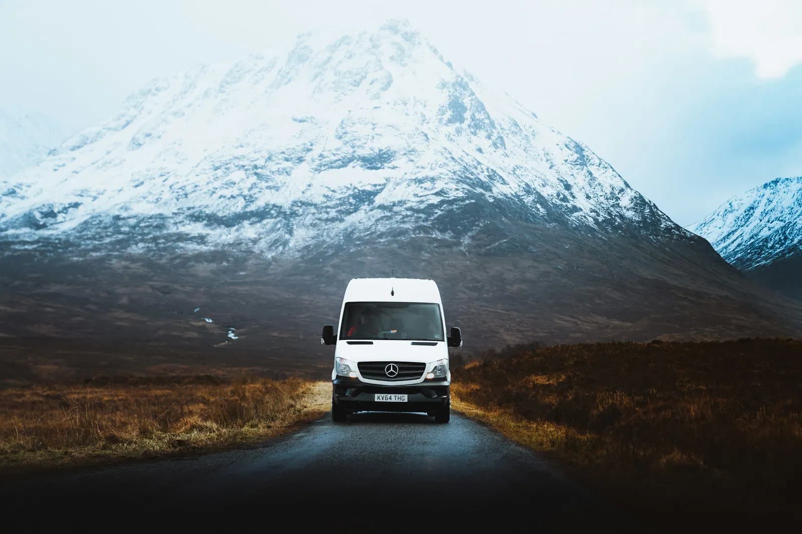 White passenger van on a narrow road leading toward a snow-capped mountain